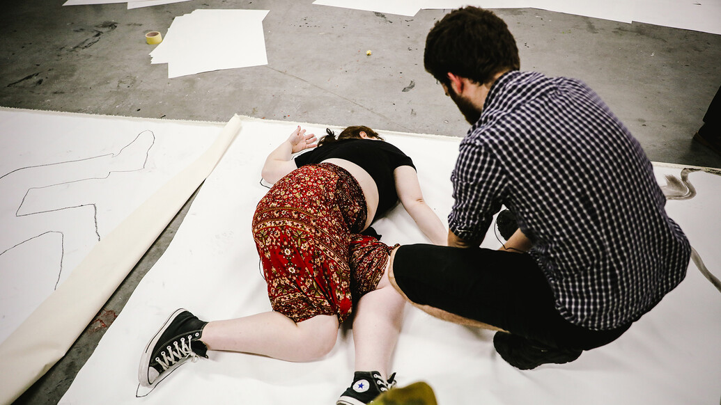A student lies down on a blank canvas whilst another examines them.