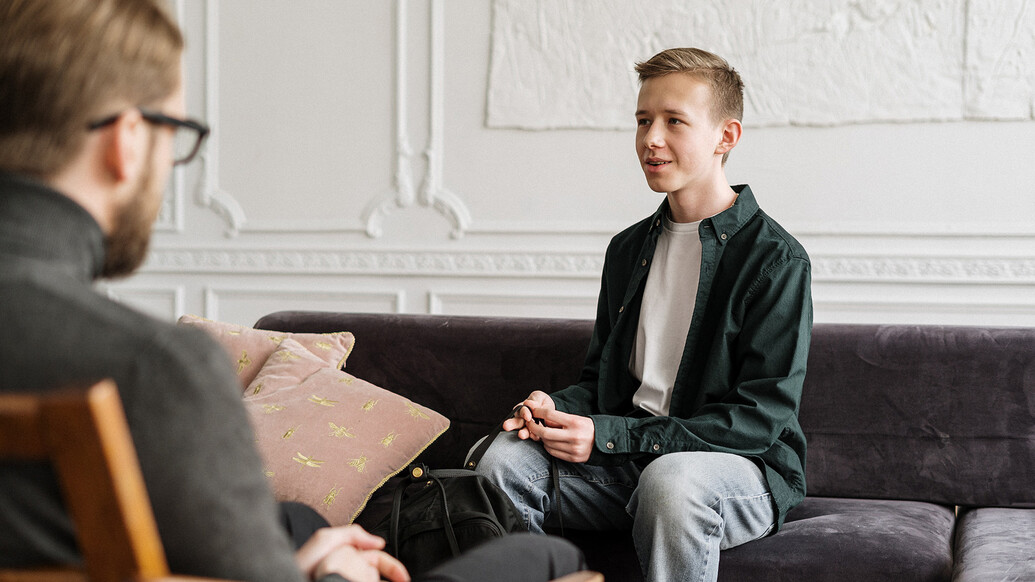 A young man sat on a sofa talking to a counsellor facing away from the camera.