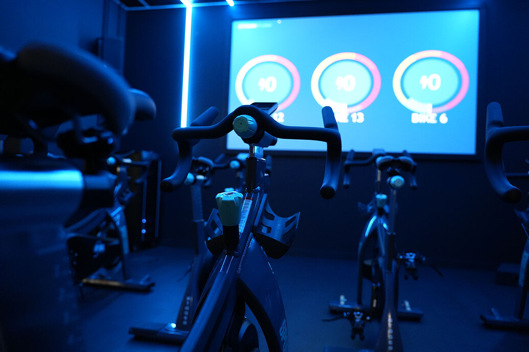 SportsDock member using the spin cycle in the virtual cycle room. On the large TV screen in front of her is an instructor.