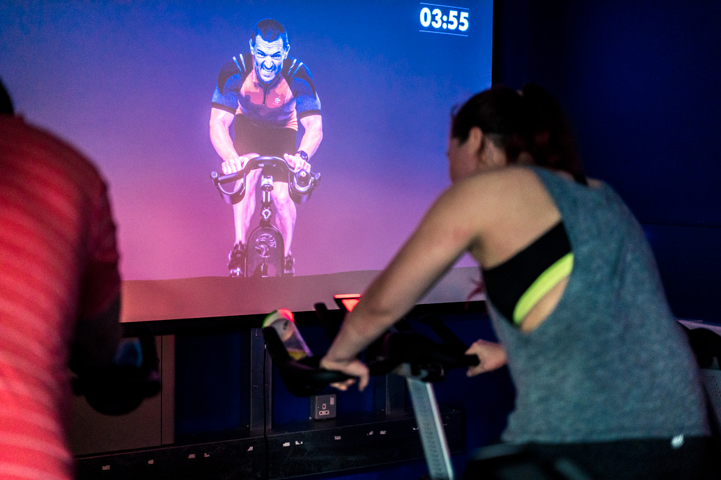 SportsDock member using teh spin syble in teh virtual cycle room. On the large TV screen in front of her is an instrucotr.
