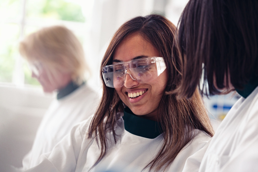 Three students in lab coats wear safety googles