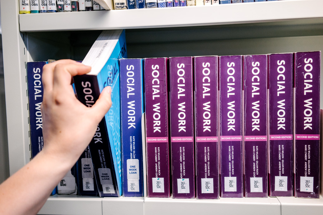 A hand pulls a book off a shelf that are lined with books in blue and purple. The books read 'SOCIAL WORK' on the spine.