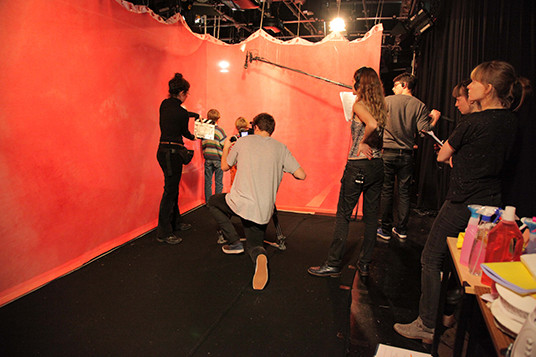 An image of a scene being filmed. Eight people stand in a room with four in the foreground and four in the foreground