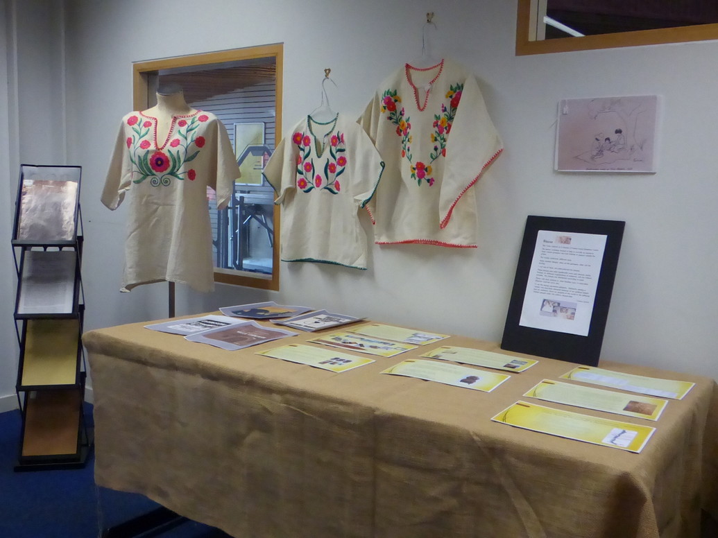 Three embroidered tunics are hanging from the wall. A display case is to the left. A table sits beneath the tunics with yellow A4 pieces of paper.