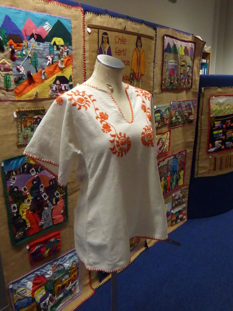An embroidered tunic sits on a mannequin. In the background, brightly coloured felt artworks are placed on a wall